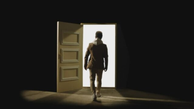 Back , rear view of man walking in front of an open door with darkness behind . Footage of person walking on floor . White light inside the door. Concept of anonymous,  spirit in coma or teleportation