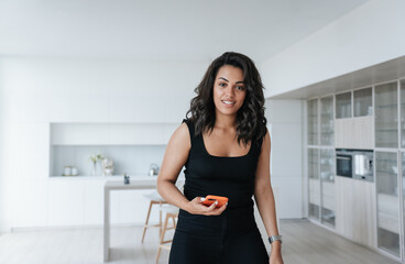 Attractive African American girl with wavy hair standing at living room holding phone looking at camera toothy smiles satisfied by new apartment. Young adult Brazilian woman enjoying weekend home.