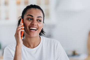 Excited African American young woman in white t-shirt talks by phone laughing looks aside preparing for party home. Adorable Brazilian girl in  good mood flirting speaks with boyfriend. Happy female