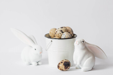 Banner. The concept of the holiday. Easter quail eggs in a white iron bucket and rabbits on a white isolated background. Beautiful Easter card.