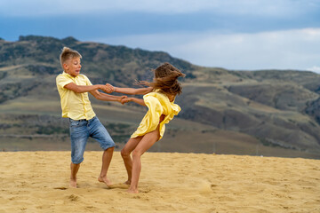 Cheerful boy and girl, holding hands, defiantly spin in sand dunes and scatter to sides with laughter and jump