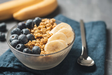 Granola cereal oatmeal with blueberries and banana fruits in a bowl on a blue napkin and grey background