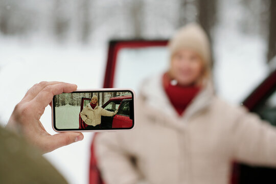 Selective focus on hand of mature man holding smartphone while taking picture of his wife in winterwear standing by open door of car