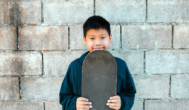 portrait of a smiling Asian kid boy holding a skateboard against the cement block wall