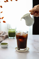 Cup of cold black coffee with a hand pouring milk into coffee glass and a cup of matcha green tea on the back in the afternoon sun on table.
Iced coffee and green matcha on a light blurred background