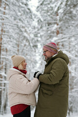Fototapeta na wymiar Smiling senior couple holding by hands and looking at one another while standing in front of camera against pinetrees covered with snow