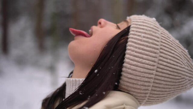 Cute girl catching snowflakes with her tongue outdoors, beige hat, Austria forest, smiling
