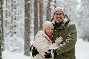 Cheerful mature couple in winterwear looking at camera during snowfall in forest covered with snow...