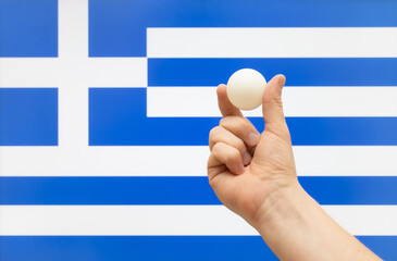 The hand holds a table tennis white ball in front of Greece flag.