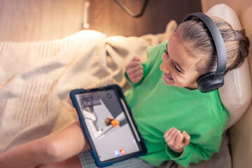 Little girl in headphones with a tablet in her room, top view.