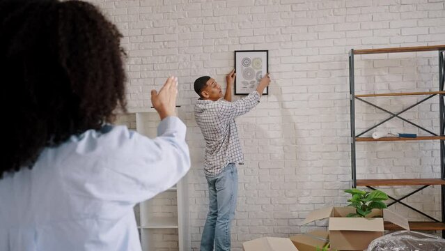 African American young man hanging a picture on the wall while moving in new apartment