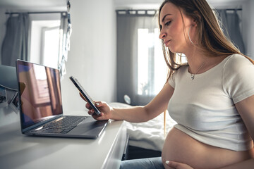 Young pregnant woman uses smartphone and laptop at home.