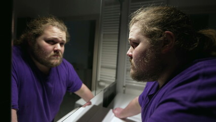 One serious overweight young man looking at bathroom mirror facing reflection. Facing fears concept