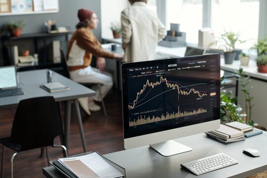 Workplace of financial analyst or trader with graphic data on screen of computer monitor standing on desk against two colleagues