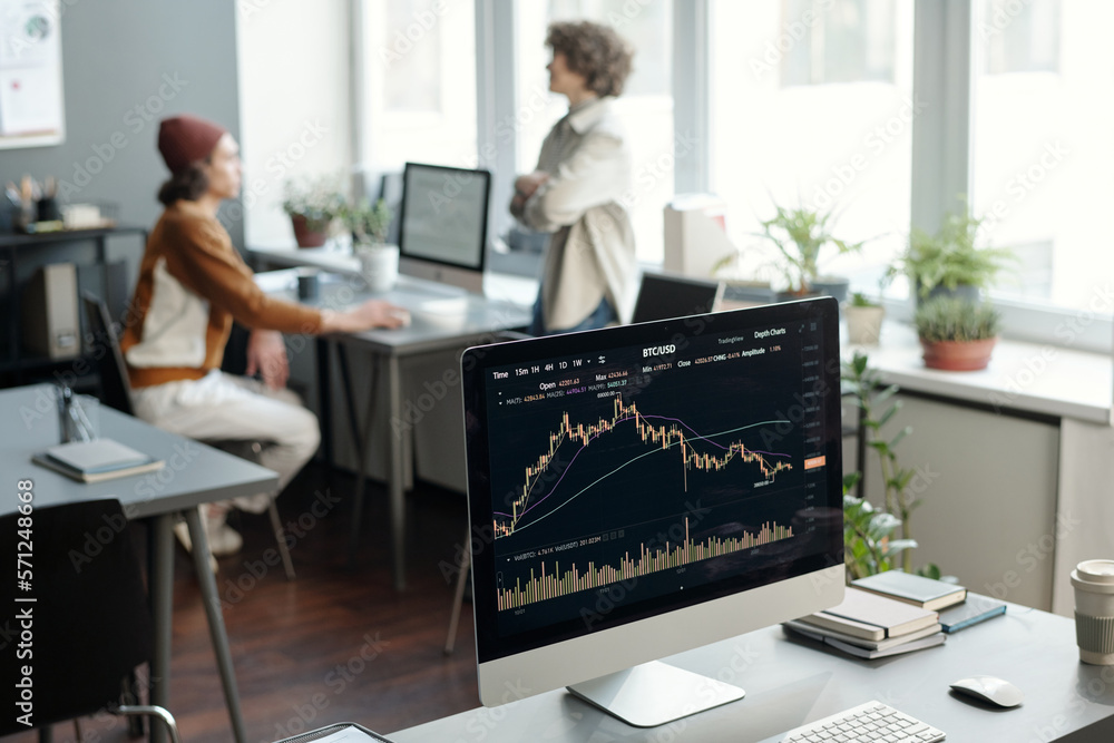 Aufkleber Computer monitor with graphic financial statistics on screen standing on workplace of broker or trader against two young economists