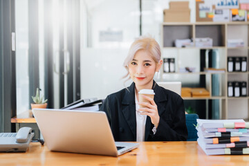 Confident beautiful Asian businesswoman typing laptop computer and digital tablet while holding coffee at office.