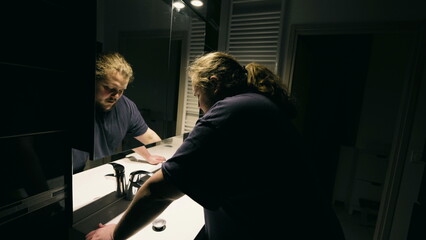 Man in emotional distress leaning on bathroom mirror suffering from mental illness alone at home....
