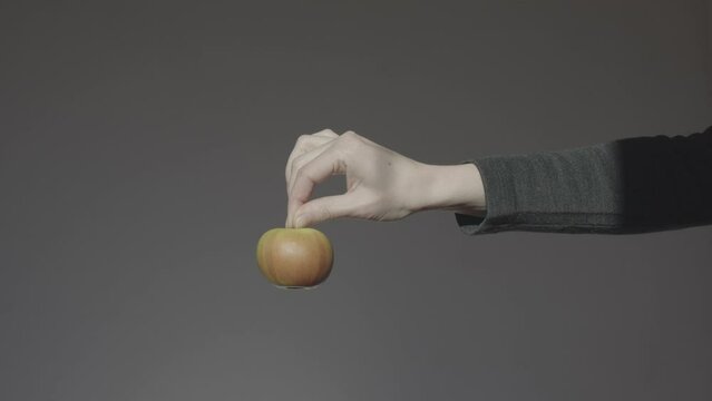 A hand on a dark background holds out a green apple holding it by the stalk. Apple of discord.