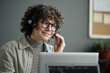 Young smiling female IT support engineer in headset looking at laptop screen and answering questions of online clients in office