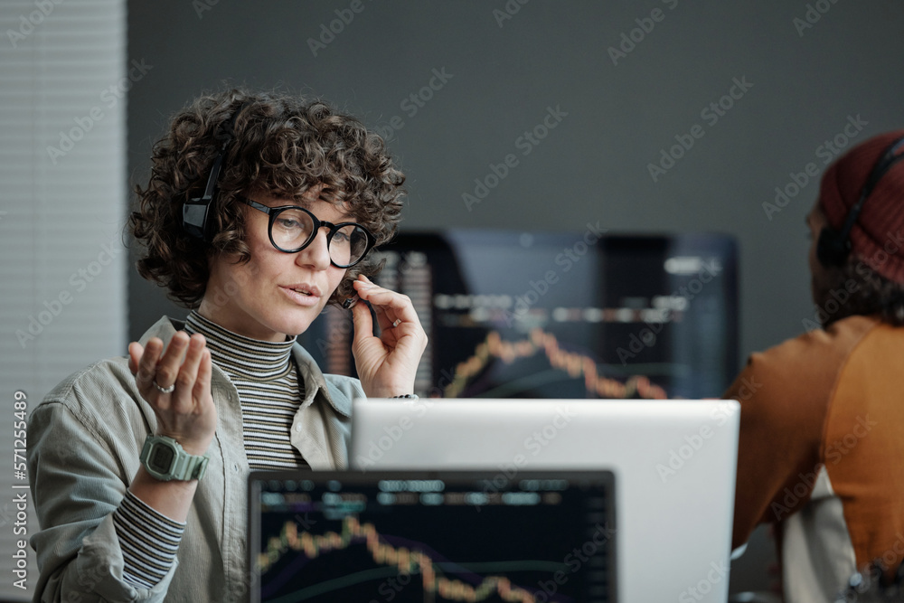 Poster Young confident operator in headset explaining something to online client while sitting by workplace in front of laptop and looking at screen