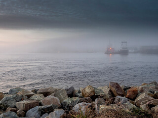 Rock coast line in foreground, ocean and background with town building and ship in fog. Calm nature scene with muted color and calm atmosphere. Galway city, Ireland. Nobody.