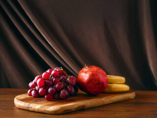 Still life with fresh ripe dark grapes, bananas and pomegranate on wooden board and table. Light...