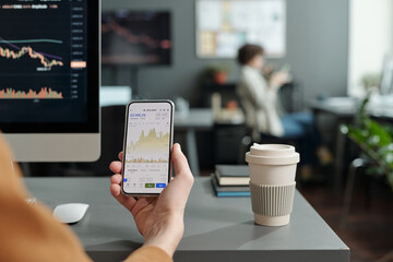 Close-up of young male economist or trader holding smartphone with graphic data on screen while sitting by workplace in office