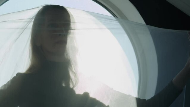 A woman in a black dress with a white transparent fabric on her head on a dark background in the rays of light from the window. Pushes fabric away from face with hands. Psychological trauma. Cocoon
