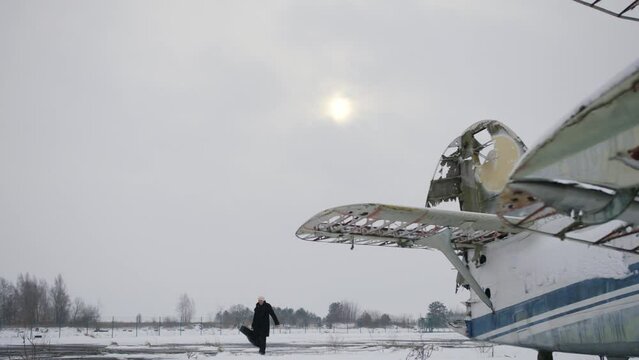 A Caucasian man in a dark coat and carrying a guitar case walks next to an old broken Soviet-made aircraft AN 2. The winter sun shines through gray clouds.