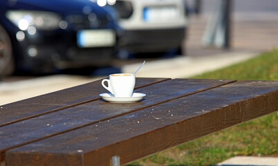 A cup of coffee on an outdoor bench, next to a car park.