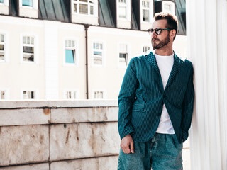 Handsome confident stylish hipster lambersexual model. Sexy modern man dressed in elegant blue suit and jeans. Fashion male posing in the street background in Europe city at sunset. In sunglasses