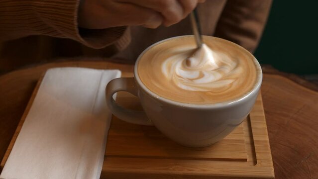 Person hand stirring latte coffee in white cup with spoon.