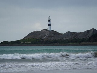Waves crashing onto ocean beach with black and white Cape Campbell lighthouse in background,...