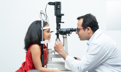 Indian middle aged professional ophthalmologist or optometrist man work at optometry clinic, using optometry instruments little girl patient to check eyesight and eyes vision at modern clinic - obrazy, fototapety, plakaty