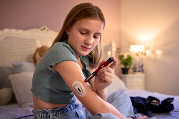 Young Diabetic Girl Sitting On Bed In Bedroom At Home Using Kit To Measure And Check Insulin Levels