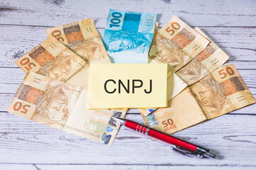 A sigla CNPJ referente ao Cadastro Nacional da Pessoa Jurídica em Português do Brasil escrita em um pedaço de papel. Notas do Real Brasileiro sobre uma mesa de madeira.
