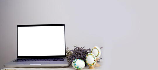 laptop with a blank screen on a table festively decorated for the Easter holiday. Adorned working place with flowers colorful eggs dried lavender flowers banner place for advertising