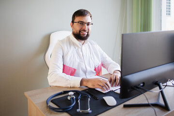 Smiling doctor looking at camera sits in an office with a computer. Doctor video conferencing, medical technology and background concept. Electronics health record system EHRs, electronics medical 