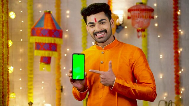 Happy smiling young man in traditional ethnic wear showing green screen mobile phone by pointing finger by looking camera - concept of app or application, promotion, festival sales and offers