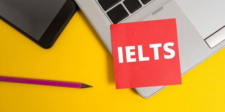 IELTS word in red sticky note paper. Laptop, smartphone, red sticky note, pencil marker, and yellow background.
