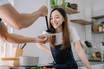 Asian young man feeding pumpkin soup to girlfriend in kitchen.