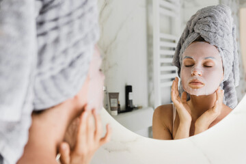 Woman in bathroom with applied sheet mask on her face looking in the mirror
