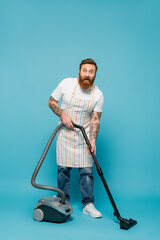 amazed tattooed man in striped apron cleaning floor with vacuum cleaner and looking at camera on blue background.