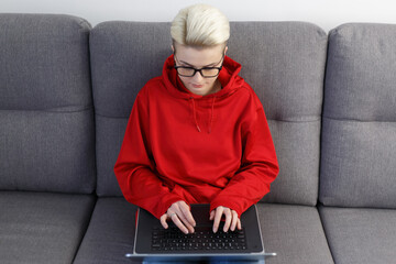 Young nerd woman in glasses sitting on couch and typing text on computer. Focused freelance person...