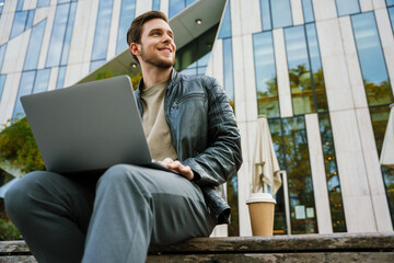 Young positive man working on laptop and drinking coffee while sitting outside