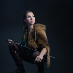 A young brave Viking heroine with a sword in hands on a dark background. A young woman in a suit in Scandinavian or fantasy style