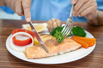 Asian senior or elderly old lady woman patient eating Salmon steak breakfast with vegetable healthy food while sitting and hungry on bed in hospital.