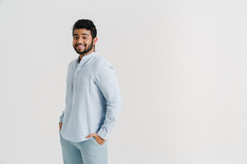 Smiling indian guy standing with hands in pockets isolated over white wall
