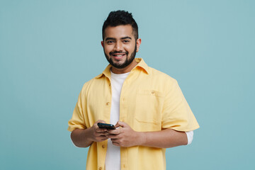Smiling indian guy using mobile phone isolated over blue wall
