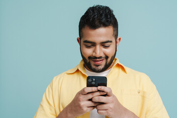 Young indian guy using mobile phone isolated over blue wall